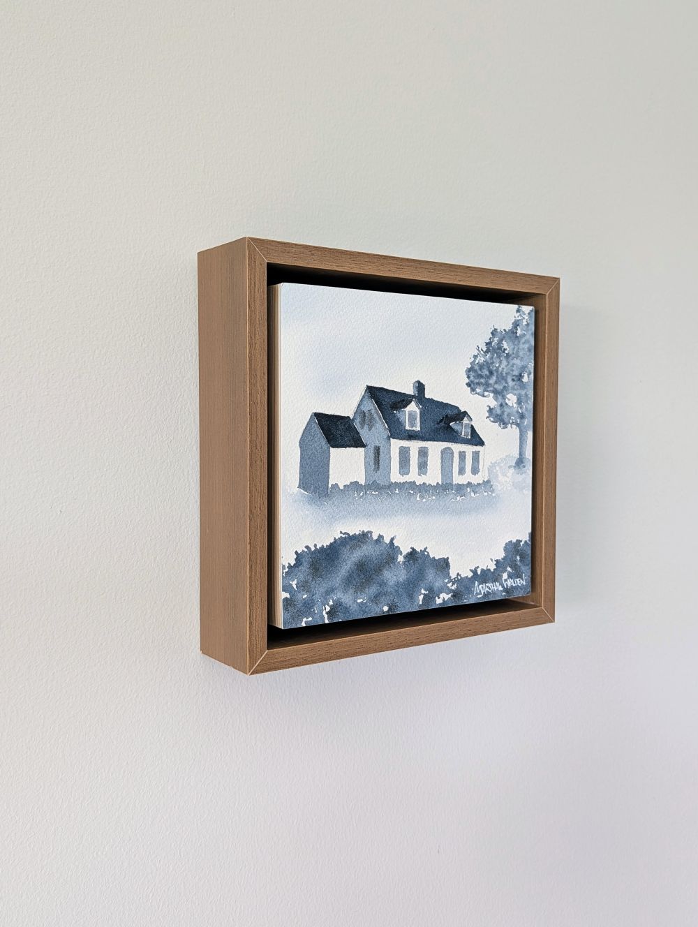 Left side view of Abide, a minimalist monochrome watercolor study of a coastal home, isolated within a deep light walnut float mount frame on a neutral wall.