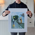 A photograph of artist Marshal Walden standing and holding up the large limited edition fine art print of his watercolor painting "Stroll In Rain." The image demonstrates the overall 16x20 scale of the piece. The print features significant extra-wide crisp white borders around the central rainy Provincetown street scene, showing the paper's rich texture and indicating its versatility for framing with or without a mat.

