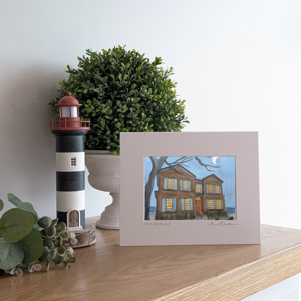 A lifestyle photograph of a white-matted watercolor print titled "After Hour Stillness" by PAAM member artist Marshal Walden, propped up on a wooden mantel. The artwork depicts the historic 2-story shingled Cape Cod home on Commercial Street made famous by the "American Horror Story: Red Tide" series. The house is shown at night with warm light glowing from its windows under a bright moon and starry sky. The print is displayed next to a black-and-white striped lighthouse figurine and a lush green potted pla
