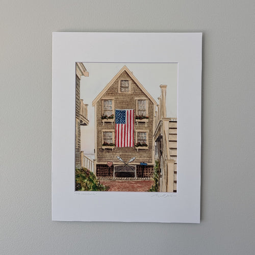 A direct overhead photograph of a white-matted watercolor print titled "77A Commercial" and signed by Marshal Walden. The print is centered within the mat, showing a shingle-style house in Provincetown with a large American flag. The entire matted artwork is centered on a plain grey wall background.
