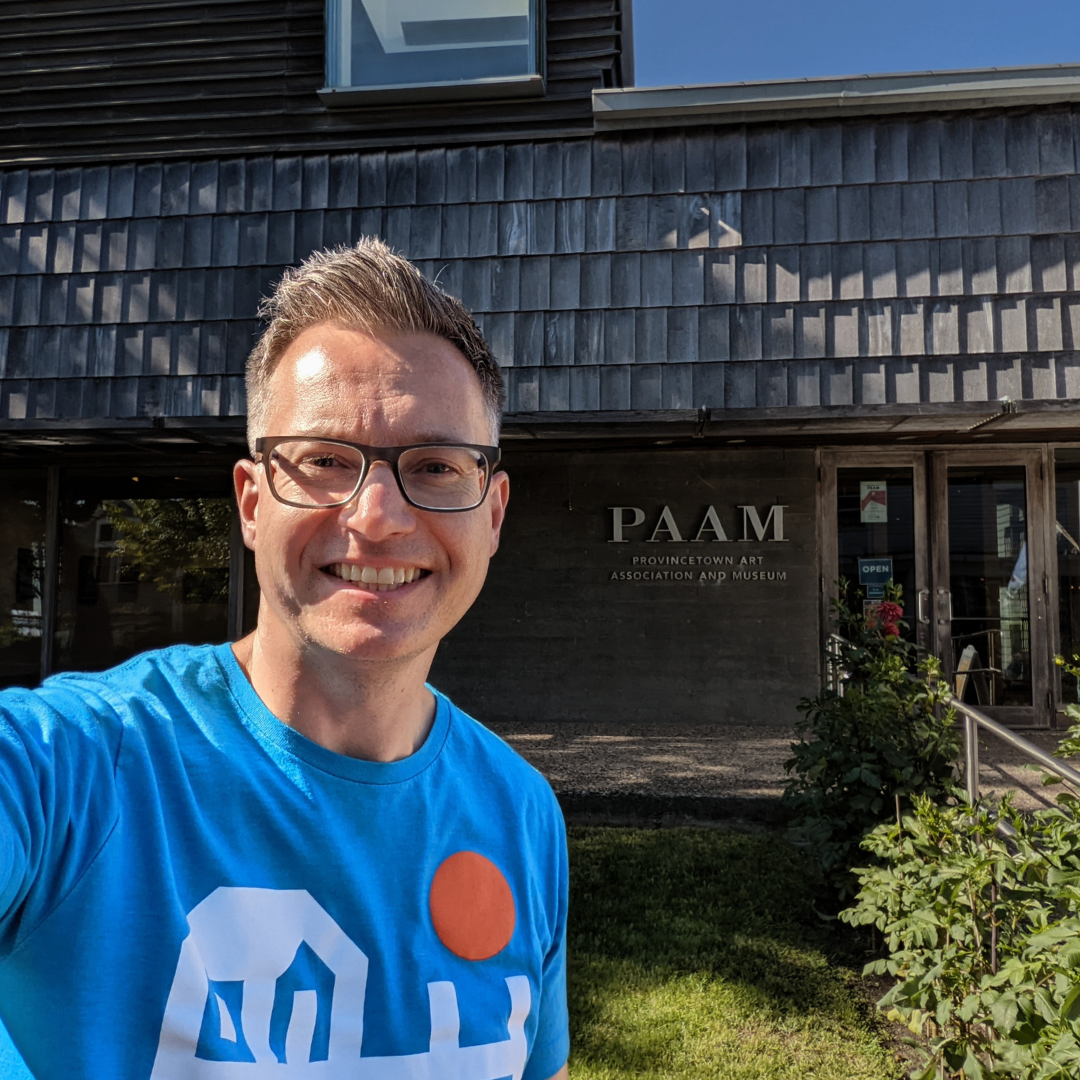 A selfie photograph taken by artist Marshal Walden, who is smiling and wearing glasses and a bright blue t-shirt. He is standing in front of the dark weathered shingle entrance of the Provincetown Art Association and Museum, with the "PAAM" sign visible on the concrete wall behind him under a sunny sky.