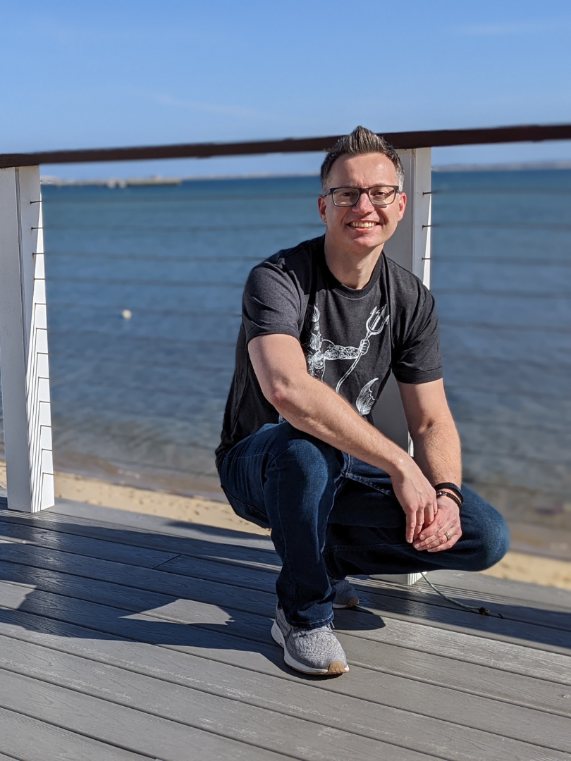 A portrait of artist Marshal Walden, crouching and smiling on a weathered wooden deck at The Red Inn. Behind him is a white railing, a sandy beach, and the expanse of Provincetown Harbor under a clear blue sky. He is wearing glasses and a dark graphic t-shirt.
