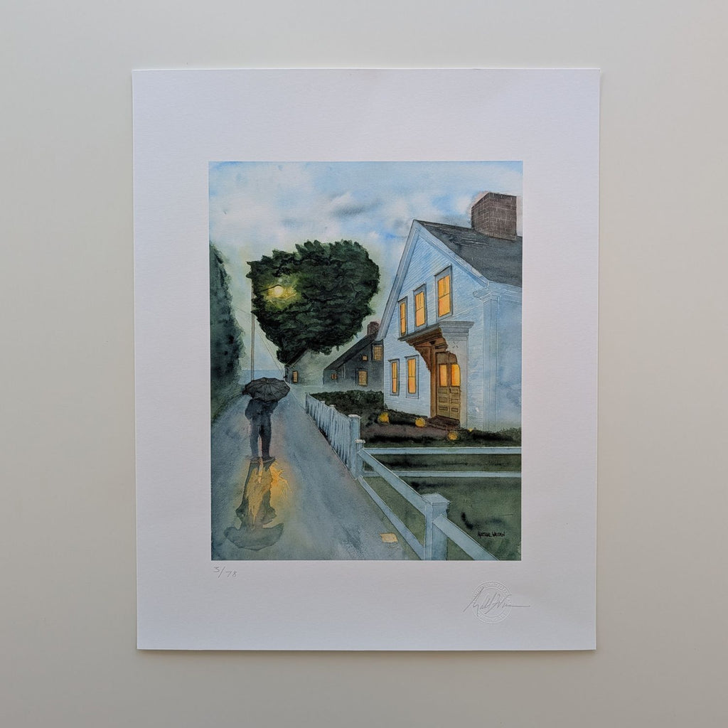 A top-down photograph of a limited edition fine art print titled "Stroll In Rain" by PAAM member artist Marshal Walden, resting flat on an off-white tabletop surface. The print is presented with a wide white border, overall 16x20. The artwork depicts a rainy early evening in Provincetown with a person walking under an umbrella, streetlights casting glistening reflections on the wet pavement, and warm glowing light emanating from a white two-story home. Visible at the bottom edge of the print are the artist'