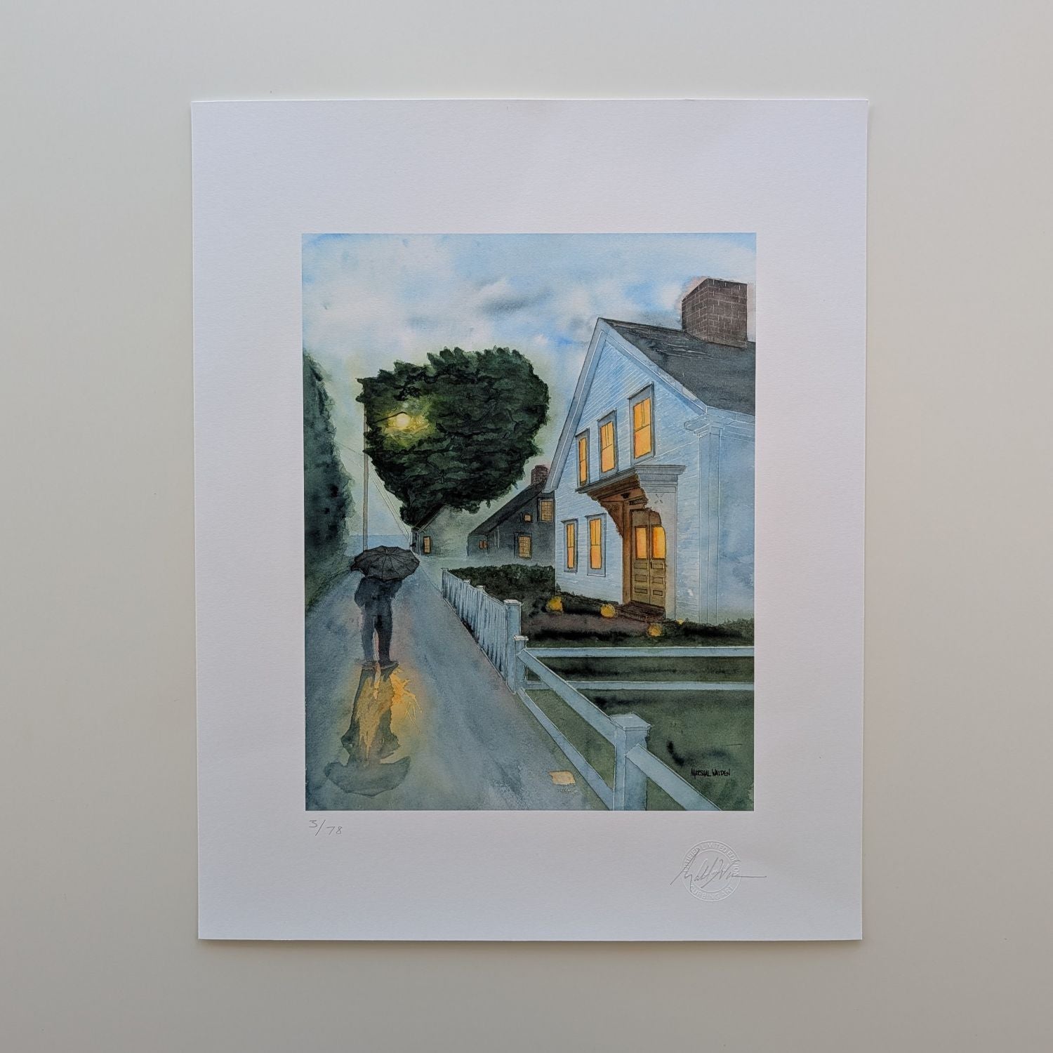 A top-down photograph of a limited edition fine art print titled "Stroll In Rain" by PAAM member artist Marshal Walden, resting flat on an off-white tabletop surface. The print is presented with a wide white border, overall 16x20. The artwork depicts a rainy early evening in Provincetown with a person walking under an umbrella, streetlights casting glistening reflections on the wet pavement, and warm glowing light emanating from a white two-story home. Visible at the bottom edge of the print are the artist'
