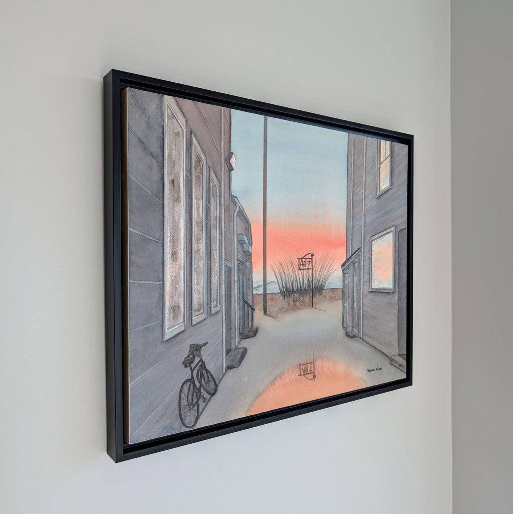 An angled side view photograph of a framed watercolor painting by PAAM member artist Marshal Walden hanging on a white wall. The perspective shows the depth of the thin black frame against the wall. The artwork inside depicts a Provincetown alley at sunrise with a bicycle and a puddle reflecting orange light.
