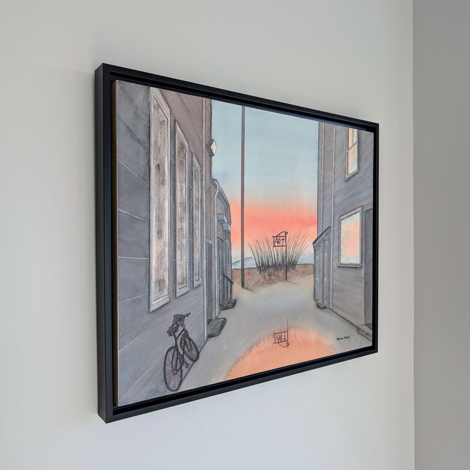 An angled side view photograph of a framed watercolor painting by PAAM member artist Marshal Walden hanging on a white wall. The perspective shows the depth of the thin black frame against the wall. The artwork inside depicts a Provincetown alley at sunrise with a bicycle and a puddle reflecting orange light.