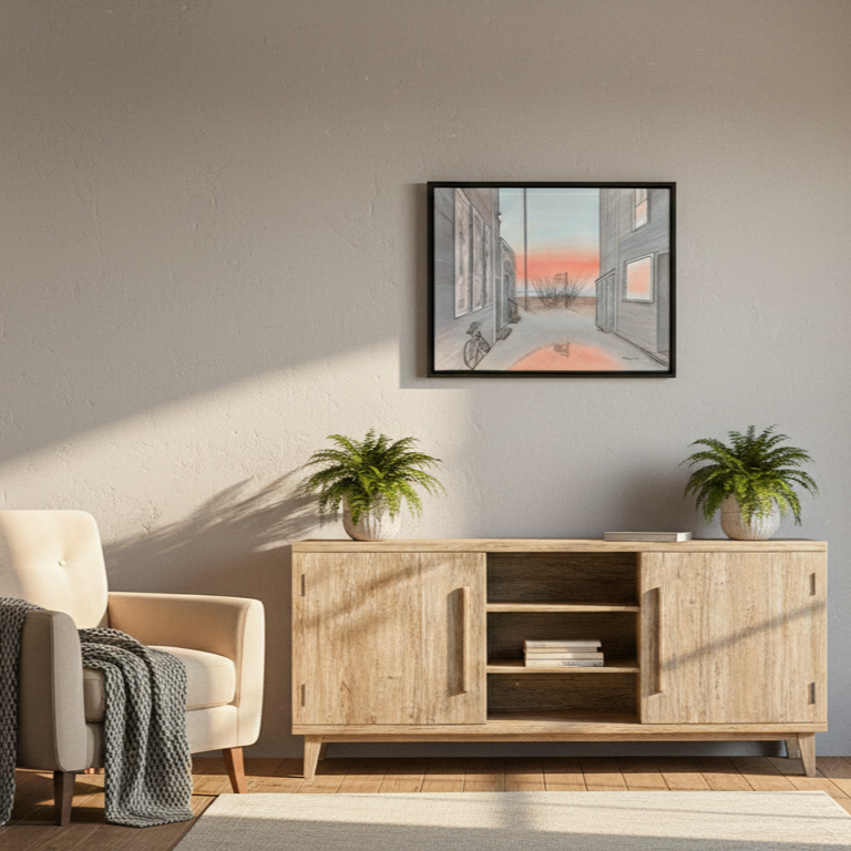 A lifestyle photograph of a black-framed watercolor painting by Marshal Walden, hanging above a light wood credenza. The painting shows a Provincetown alley at sunrise with a bicycle and a reflecting puddle. The room features a beige armchair with a grey blanket, two potted ferns, books, and warm, directional sunlight creating long shadows on the wall.