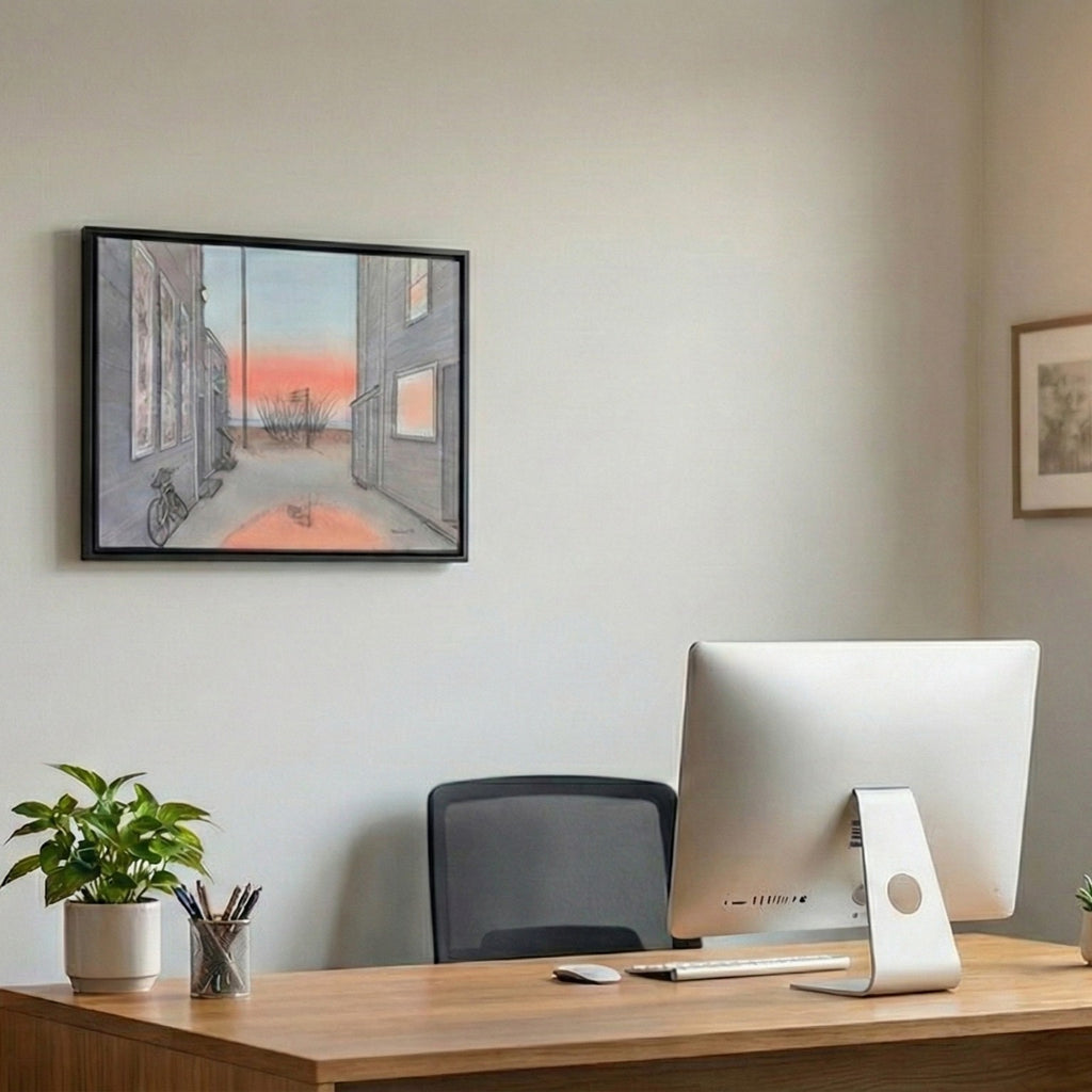 A lifestyle photograph of a black-framed watercolor painting by PAAM member artist Marshal Walden, hanging on a white wall in a home office. The painting shows a Provincetown alley at sunrise. Below it is a wooden desk with a large computer monitor, a potted plant, and a pen holder.