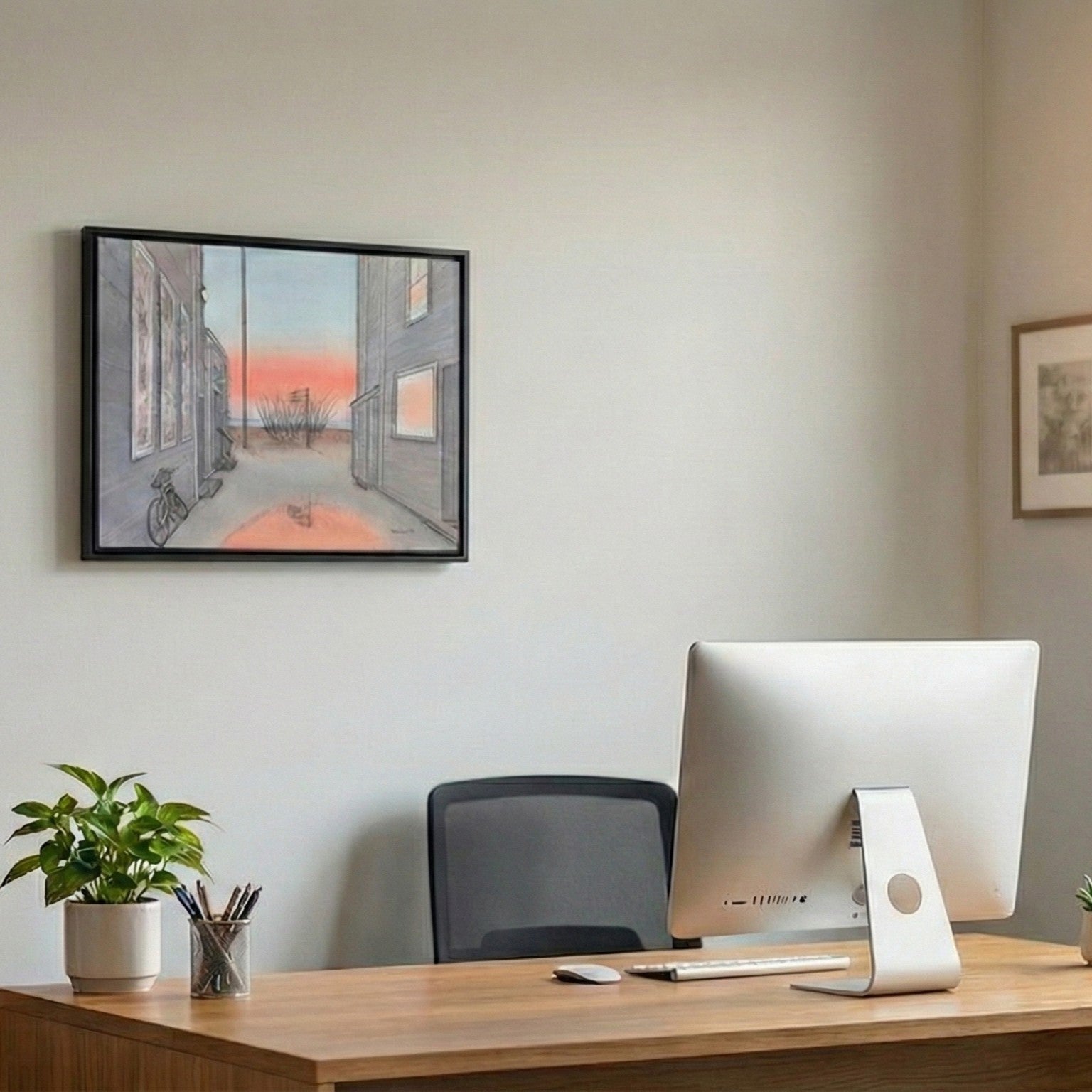 A lifestyle photograph of a black-framed watercolor painting by PAAM member artist Marshal Walden, hanging on a white wall in a home office. The painting shows a Provincetown alley at sunrise. Below it is a wooden desk with a large computer monitor, a potted plant, and a pen holder.