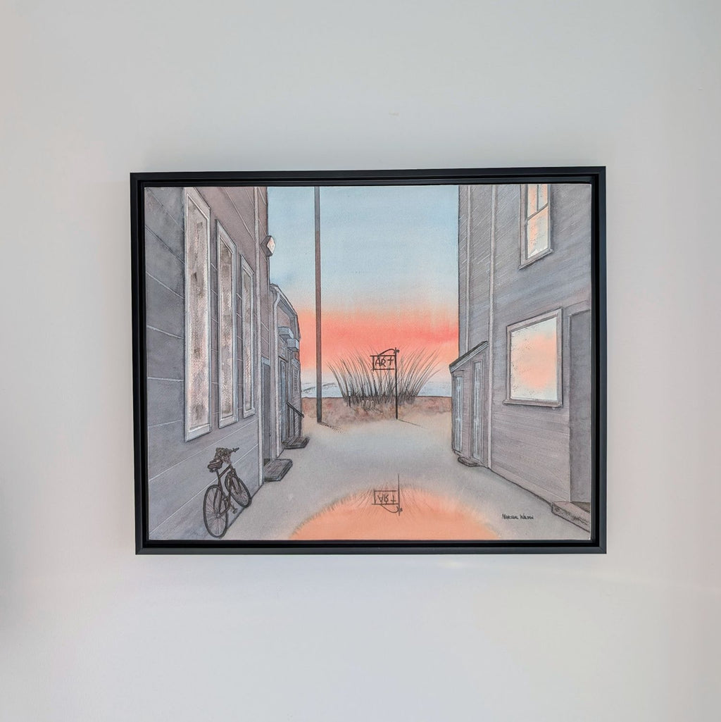 A photograph of a black-framed watercolor painting by PAAM member artist Marshal Walden, hanging centered on a plain light grey wall. The painting depicts a narrow Provincetown alley at sunrise, with a bicycle on the left, an "ART" sign by the beach in the distance, and a puddle reflecting the vibrant orange and pink sky. Signed "MARSHAL WALDEN" in the bottom right corner of the painting.