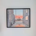 A photograph of a black-framed watercolor painting by PAAM member artist Marshal Walden, hanging centered on a plain light grey wall. The painting depicts a narrow Provincetown alley at sunrise, with a bicycle on the left, an "ART" sign by the beach in the distance, and a puddle reflecting the vibrant orange and pink sky. Signed "MARSHAL WALDEN" in the bottom right corner of the painting.