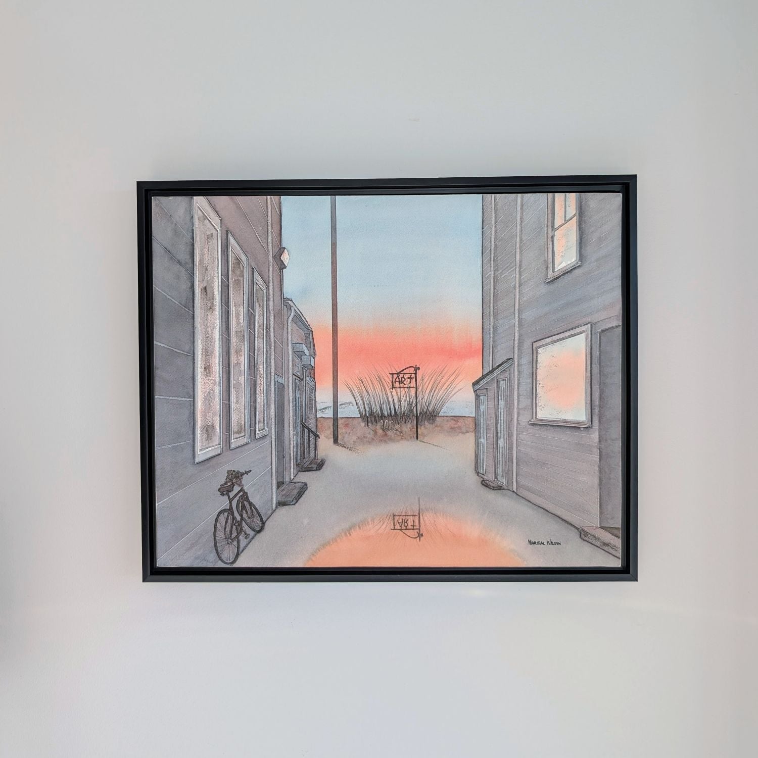 A photograph of a black-framed watercolor painting by PAAM member artist Marshal Walden, hanging centered on a plain light grey wall. The painting depicts a narrow Provincetown alley at sunrise, with a bicycle on the left, an "ART" sign by the beach in the distance, and a puddle reflecting the vibrant orange and pink sky. Signed "MARSHAL WALDEN" in the bottom right corner of the painting.