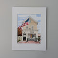 A professional open edition watercolor print titled "Firehouse Pride" with matting titled and signed by PAAM member artist Marshal Walden. The painting depicts the historic Engine Co. 1 firehouse in Provincetown on a bright, sunny day, featuring a large American flag waving prominently and a Pride flag displayed in the open bay porch. The artwork is presented in a crisp white mat and centered against a neutral beige wall.
