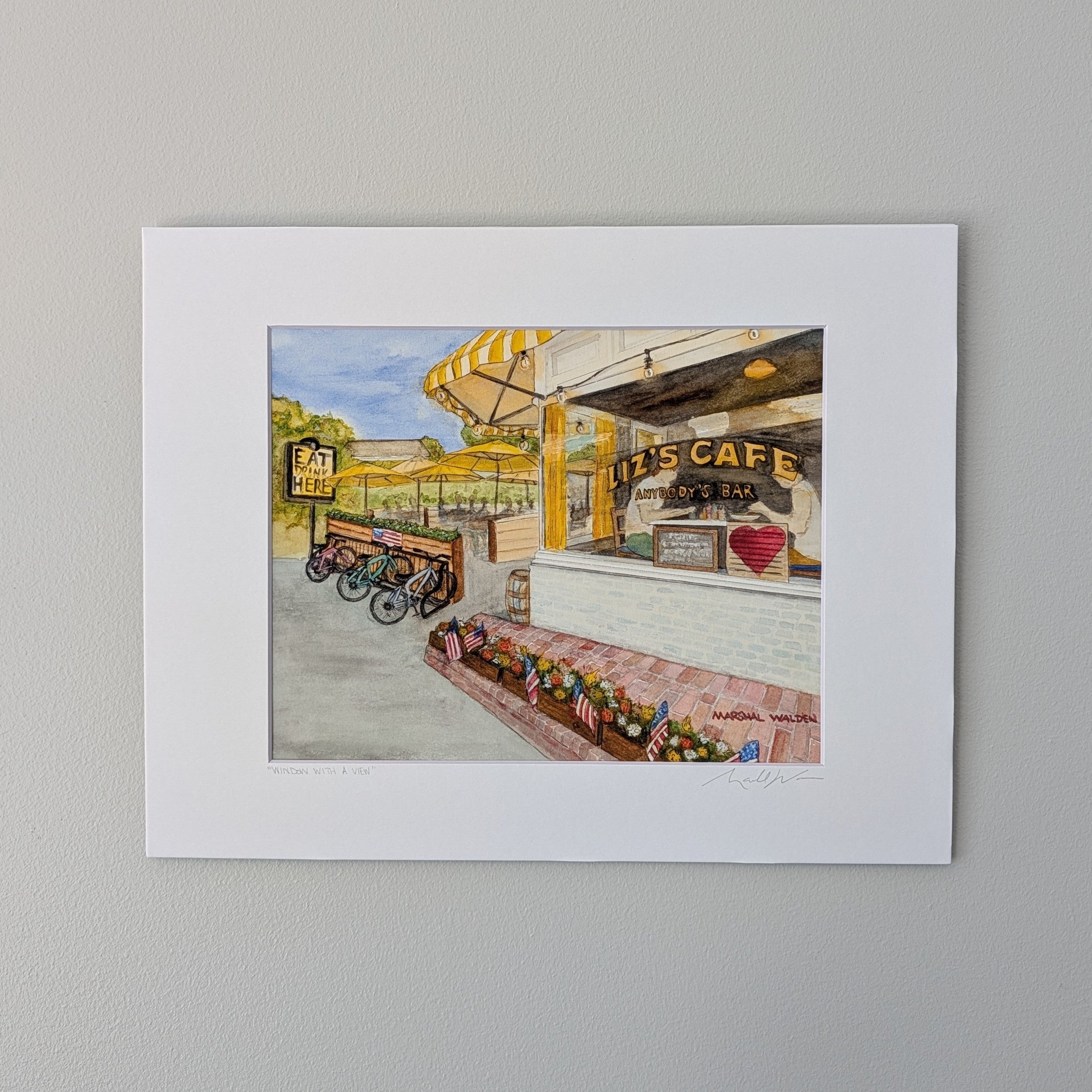 A photograph of a matted watercolor print titled "Window With A View" with titled and signed matting by Marshal Walden, displayed against a light gray wall. The print depicts the Liz's Café Anybody's Bar with the iconic yellow umbrellas, bright yellow sign, and big picture windows in the front along Bradford Street in Provincetown.
