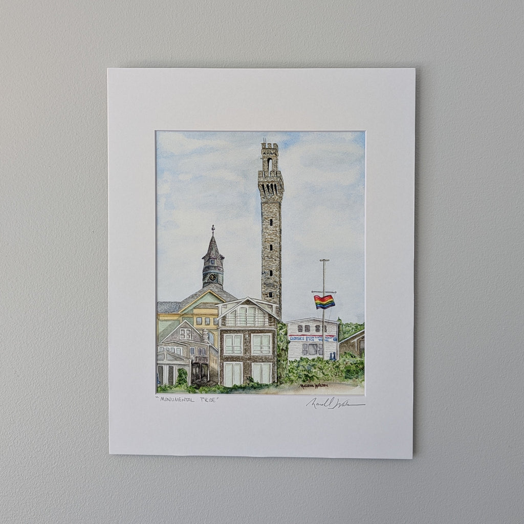 A professionally printed fine art watercolor print titled "Monumental Pride", hand-titled and signed by PAAM member artist Marshal Walden on the matting. The artwork depicts the historic skyline of Provincetown, featuring the tall granite Pilgrim Monument rising prominently above the town hall clock tower, local businesses, and coastal residences under a cloudy blue sky. A rainbow Pride flag waves on a flagpole to the right. The print is displayed in a crisp white mat against a neutral beige wall.

