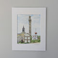 A professionally printed fine art watercolor print titled "Monumental Pride", hand-titled and signed by PAAM member artist Marshal Walden on the matting. The artwork depicts the historic skyline of Provincetown, featuring the tall granite Pilgrim Monument rising prominently above the town hall clock tower, local businesses, and coastal residences under a cloudy blue sky. A rainbow Pride flag waves on a flagpole to the right. The print is displayed in a crisp white mat against a neutral beige wall.
