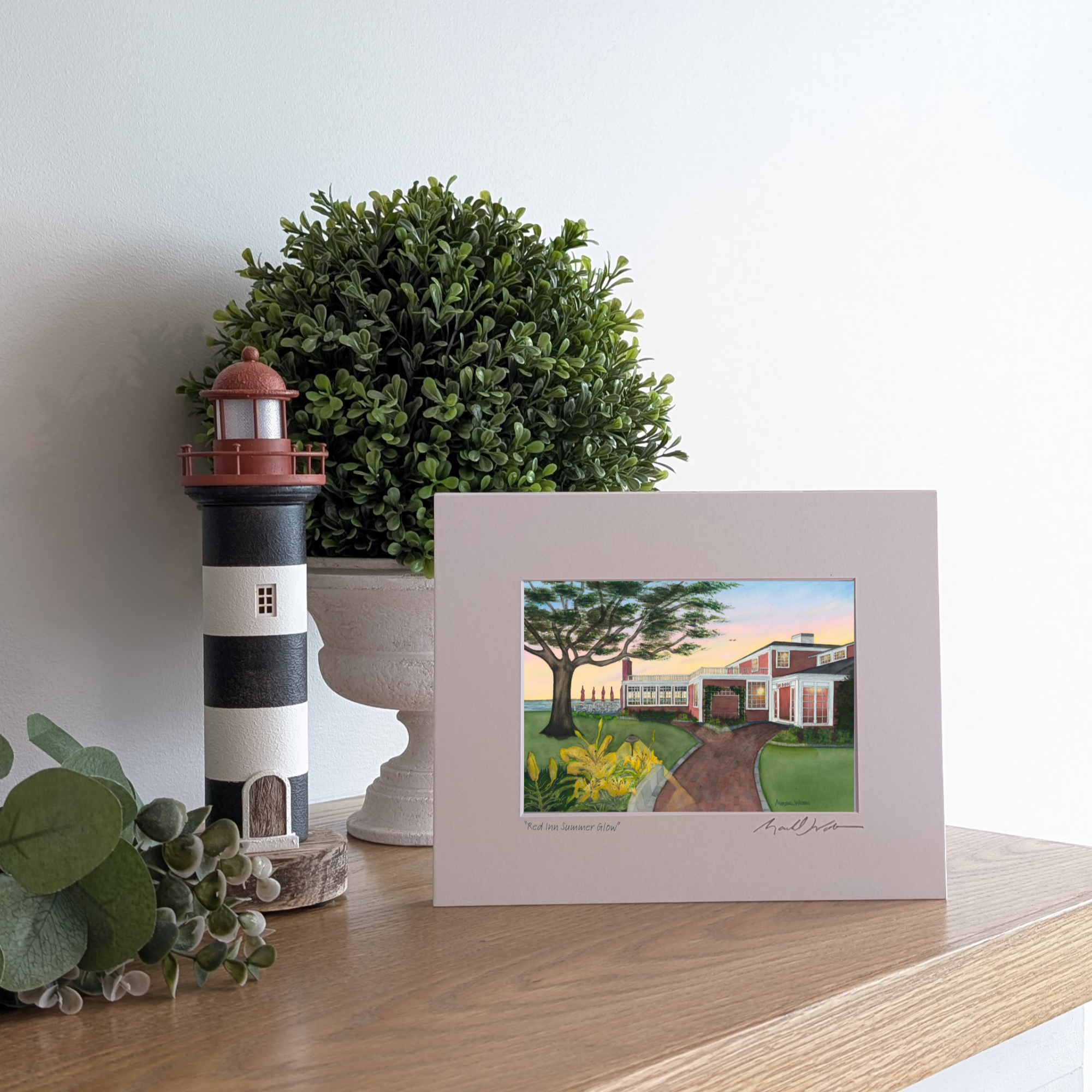 A lifestyle photograph of a professionally printed watercolor open edition titled "Red Inn Summer Glow" and signed by artist Marshal Walden. The artwork captures the iconic building at sunset with long casting shadows. The print is propped up on a light wood mantel next to a black-and-white striped lighthouse figurine and a lush green boxwood plant.
