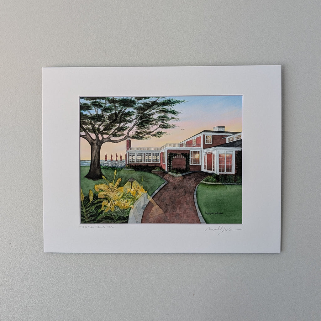 A professionally printed fine art watercolor open edition titled "Red Inn Summer Glow", hand-titled and signed by artist Marshal Walden, centered against a neutral beige wall. The artwork captures the iconic Provincetown building bathed in warm summer sunset light, casting long, deep shadows that emphasize its architectural form.
