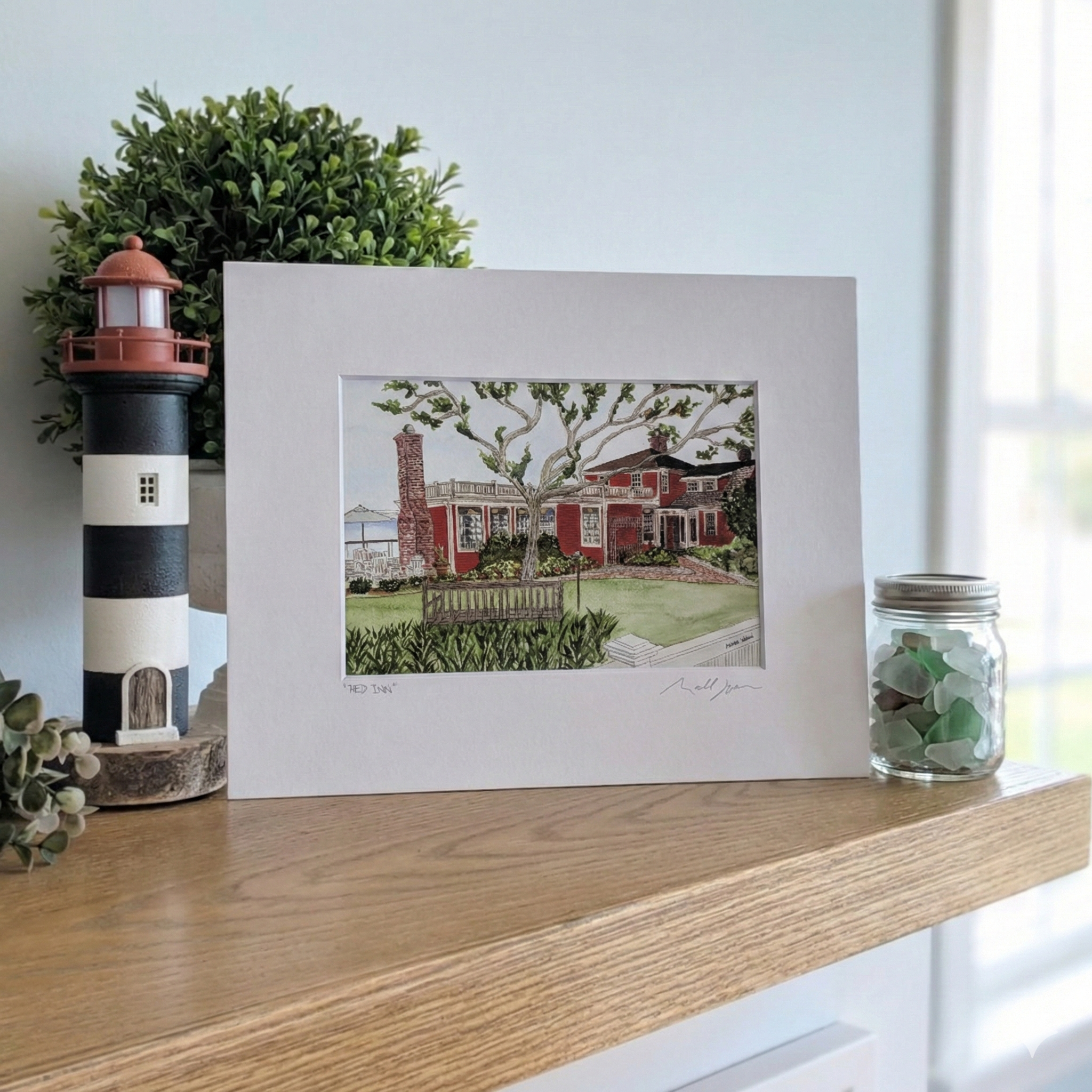 A lifestyle photograph of a professionally printed watercolor print titled "Red Inn" by PAAM artist Marshal Walden, propped up on a wooden mantel. The artwork depicts the iconic red building in Provincetown, distilled to its architectural bones. Next to the print is a black and white striped lighthouse figurine, lush green plant, and a clear glass jar filled with green and white sea glass.
