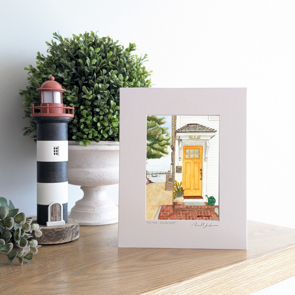 A photograph of a matted art print titled "The Turn - Yellow Door," propped up on a wooden mantel. The artwork depicts a bright yellow door on a white coastal home in Provincetown, with colorful kayaks and a sandy beach path leading toward the harbor and a distant pier. To the left of the print are a black-and-white striped lighthouse figurine and a lush green potted plant.
