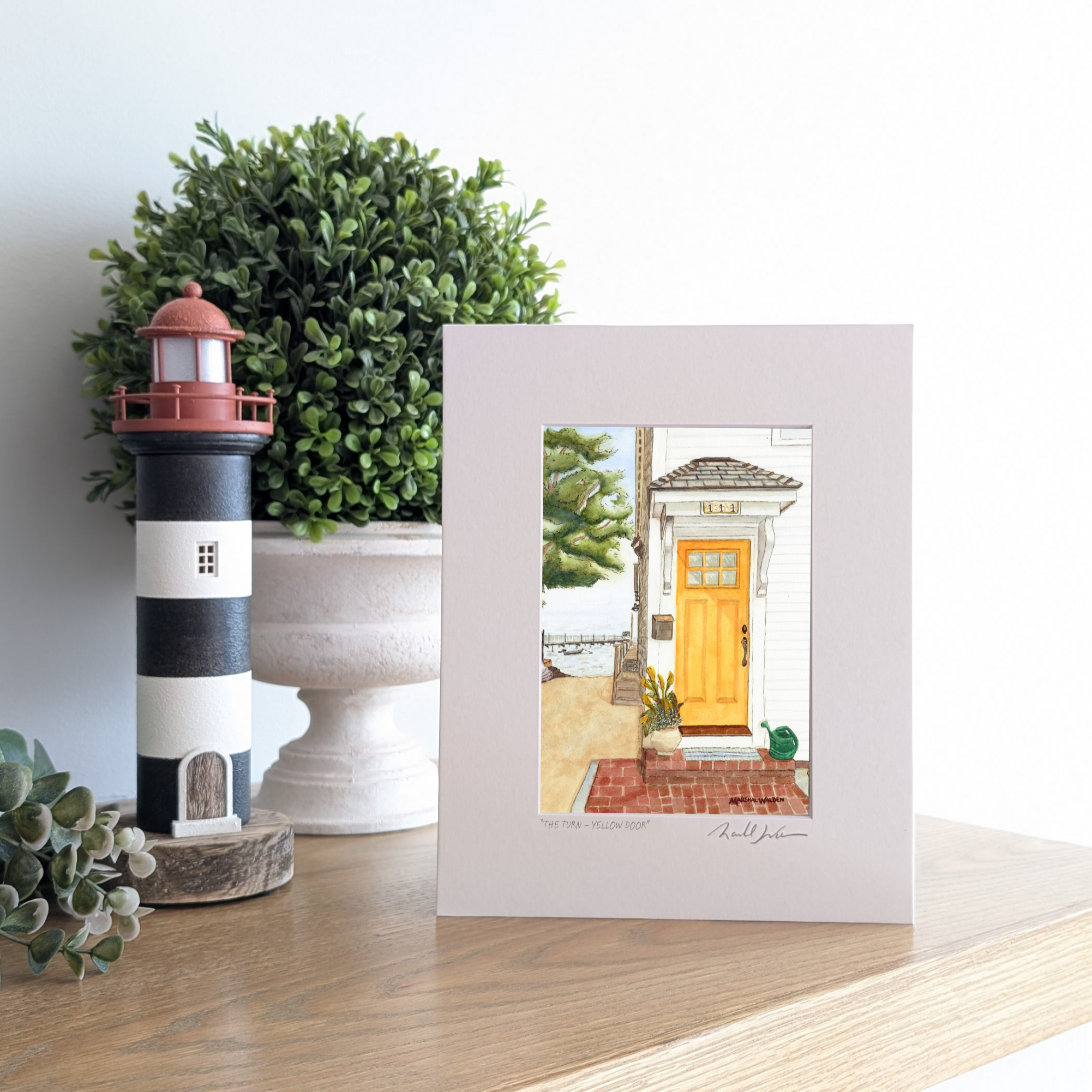 A photograph of a matted art print titled "The Turn - Yellow Door," propped up on a wooden mantel. The artwork depicts a bright yellow door on a white coastal home in Provincetown, with colorful kayaks and a sandy beach path leading toward the harbor and a distant pier. To the left of the print are a black-and-white striped lighthouse figurine and a lush green potted plant.

