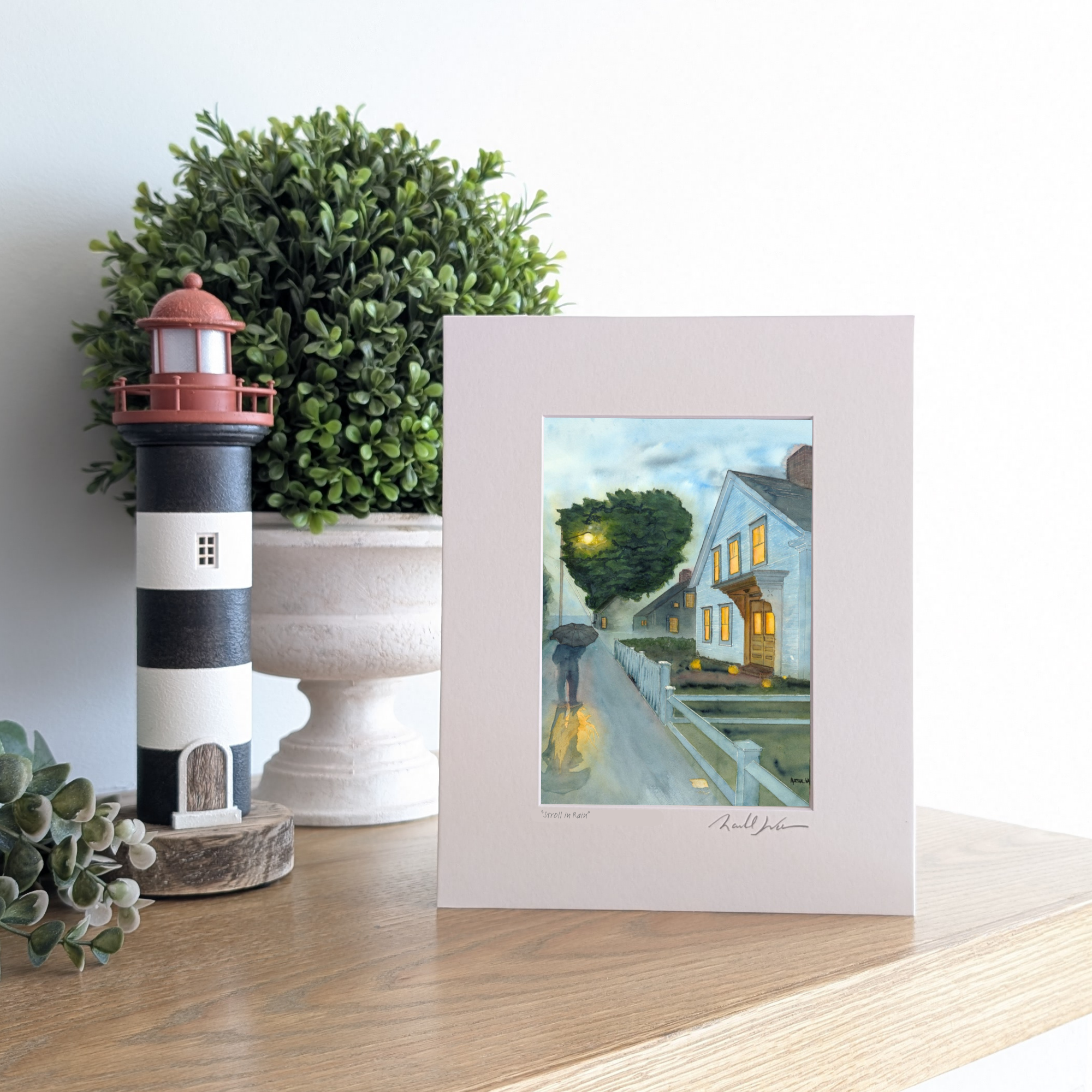 A lifestyle photograph of a professionally printed watercolor open edition titled "Stroll In Rain" by PAAM member artist Marshal Walden, propped up on a wooden mantel. The artwork depicts a rainy early evening in Provincetown with a person walking under an umbrella, streetlamp reflections on wet pavement, and warm light glowing from a white two-story home. The print sits next to a black-and-white striped lighthouse figurine and a lush green plant.

