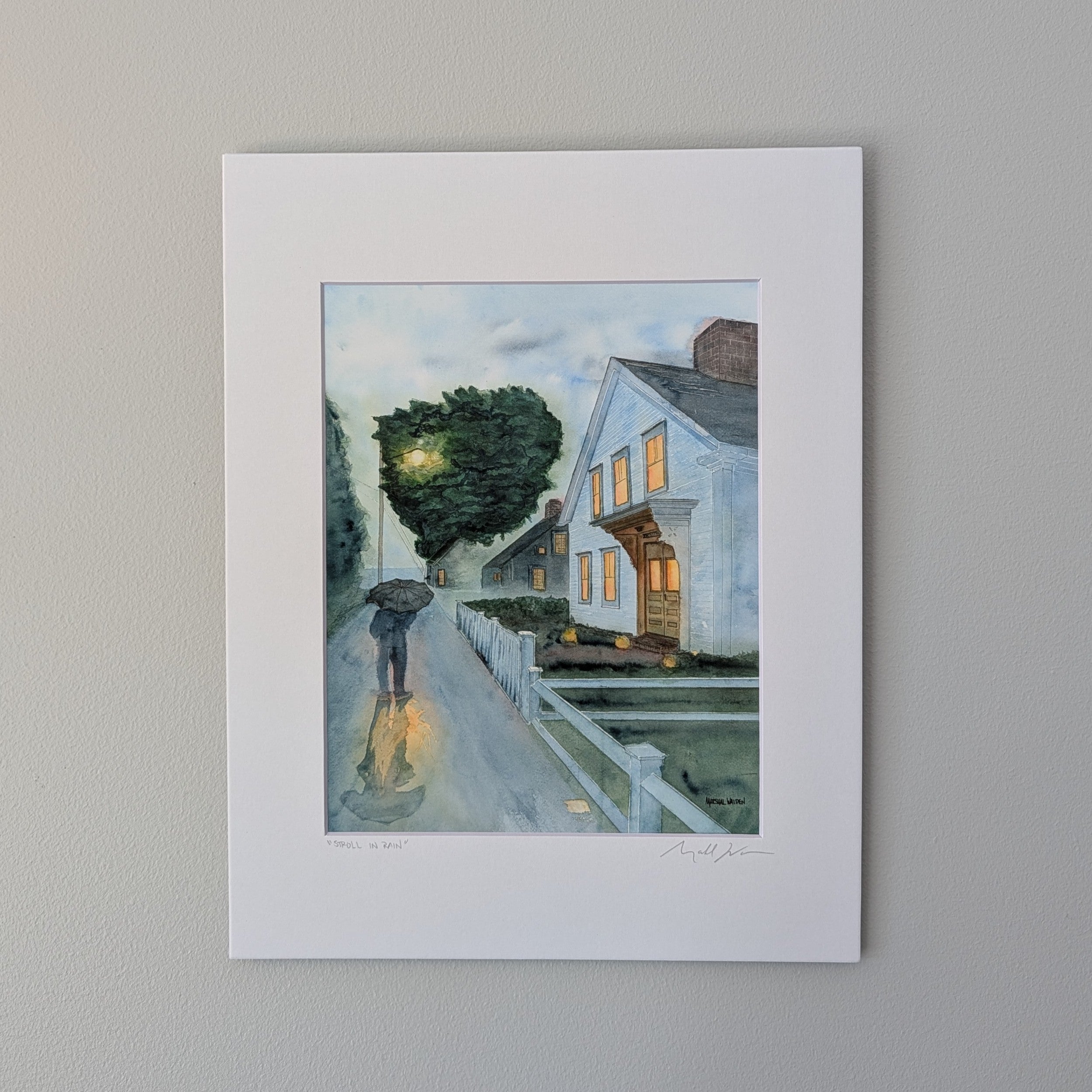 A professional fine art watercolor print titled "Stroll In Rain", hand-titled and signed by PAAM member artist Marshal Walden. The artwork depicts a rainy early evening on a Provincetown street where a person walks under a black umbrella. Warm light glows from the windows of a white two-story home, and a streetlamp creates glistening reflections on the wet pavement. The piece is presented in a crisp white mat and displayed against a light beige wall.
