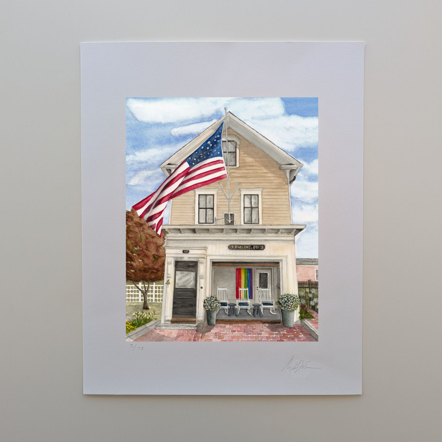 A photograph of a 16x20 limited edition fine art print titled "Firehouse Pride" by PAAM member artist Marshal Walden, resting on an off-white tabletop. The watercolor artwork depicts the historic Engine Co. 1 firehouse in Provincetown on a sunny day, featuring a large American flag waving in the breeze and a rainbow Pride flag displayed in the open bay porch. The artist's hand-penciled signature, the edition number out of 78, and a raised Curbing Art embossed seal are visible on the bottom border.
