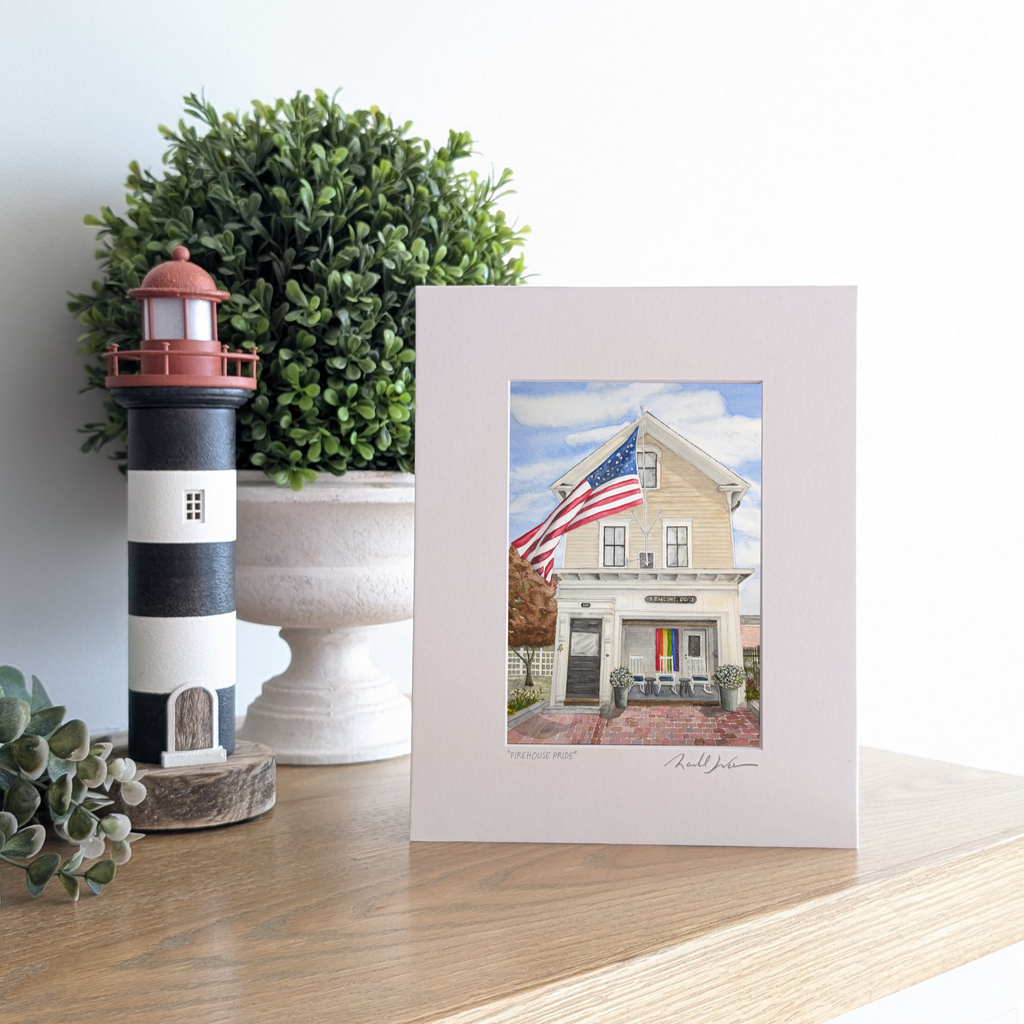 A lifestyle photograph of a professionally printed watercolor print titled "Firehouse Pride" by PAAM member artist Marshal Walden, propped up on a wooden mantel. The artwork depicts the historic Engine Co. 1 firehouse in Provincetown on a bright sunny day, featuring a large American flag waving and a rainbow Pride flag displayed in the open bay porch. The print sits next to a black and white striped lighthouse figurine and a lush boxwood plant.
