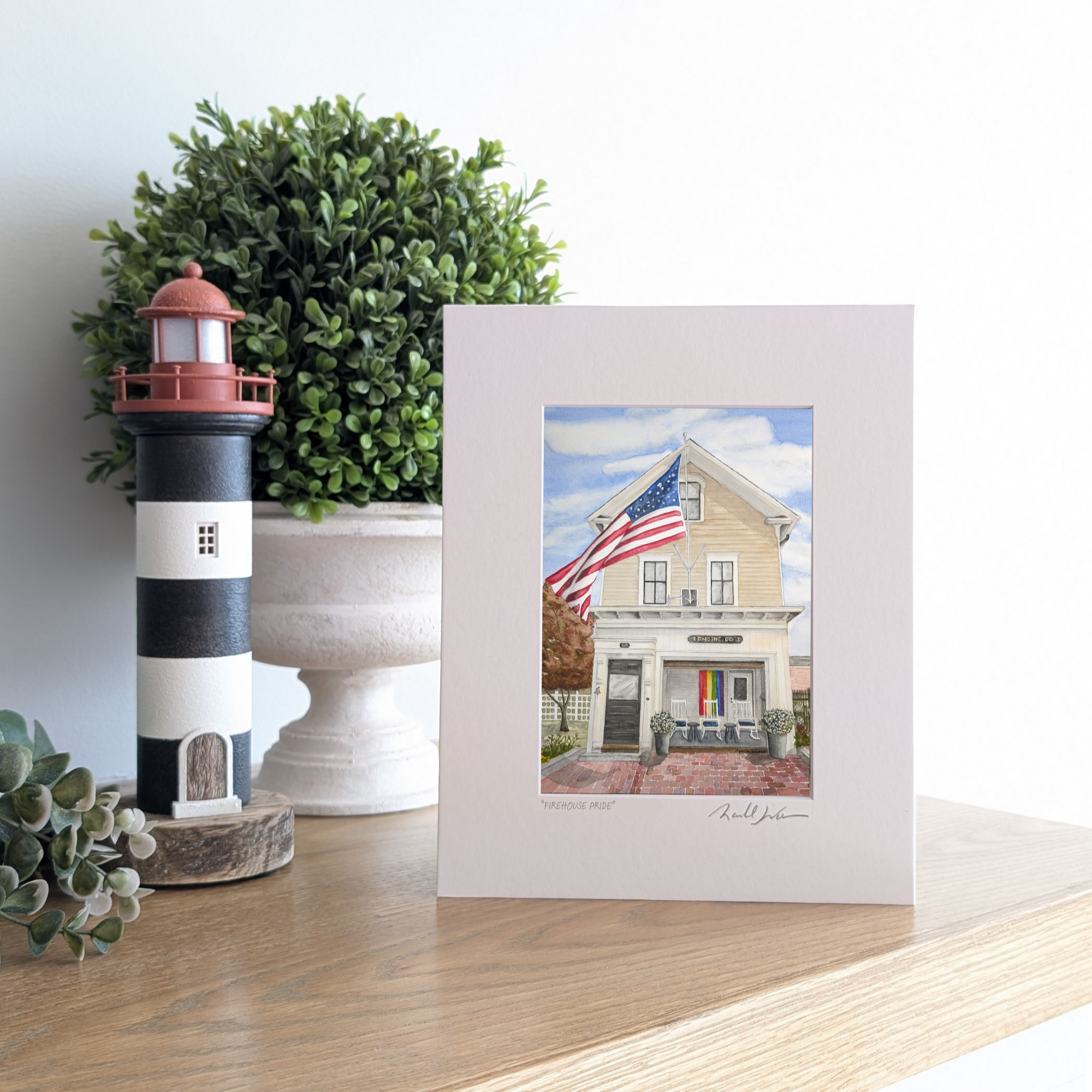A lifestyle photograph of a professionally printed watercolor print titled "Firehouse Pride" by PAAM member artist Marshal Walden, propped up on a wooden mantel. The artwork depicts the historic Engine Co. 1 firehouse in Provincetown on a bright sunny day, featuring a large American flag waving and a rainbow Pride flag displayed in the open bay porch. The print sits next to a black and white striped lighthouse figurine and a lush boxwood plant.
