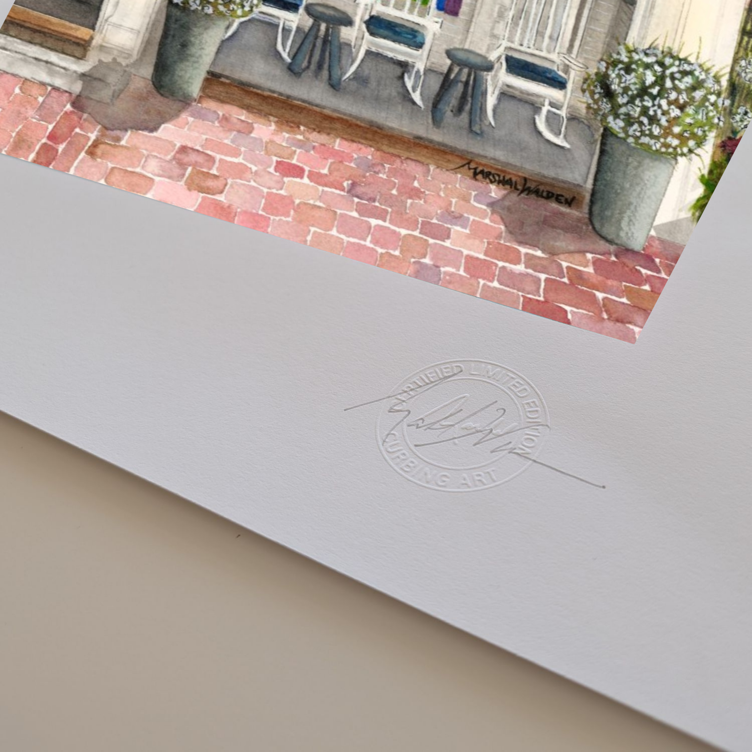 A macro photograph showing the lower corner details of the limited edition fine art print "Firehouse Pride." The focus is on artist Marshal Walden's signature written in graphite pencil next to the raised, circular official Curbing Art studio embossing seal pressed into the surface. The lighting and close perspective emphasize the deep texture, tooth, and substantial thickness of the heavy archival paper stock.

