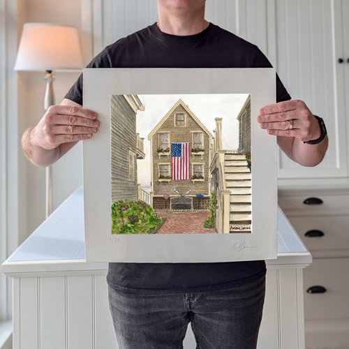 A photograph of artist Marshal Walden holding the 16x16 limited edition fine art print "77A Commercial" in the studio. The image demonstrates the print's scale and highlights the detailed watercolor texture and extra-wide crisp white borders, which allow for versatile framing either without a mat for a modern look or matted to a standard 10x10 opening for a traditional presentation. His pencil signature and edition numbering are visible on the bottom border.

