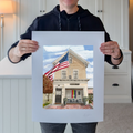 A photograph of artist Marshal Walden standing and holding up the large 16x20 limited edition fine art print of his painting "Firehouse Pride." The image demonstrates the scale of the piece, which features the historic Provincetown firehouse with American and Pride flags. The print has significant extra-wide crisp white borders, showing the paper's texture and indicating its versatility for framing with or without a mat.
