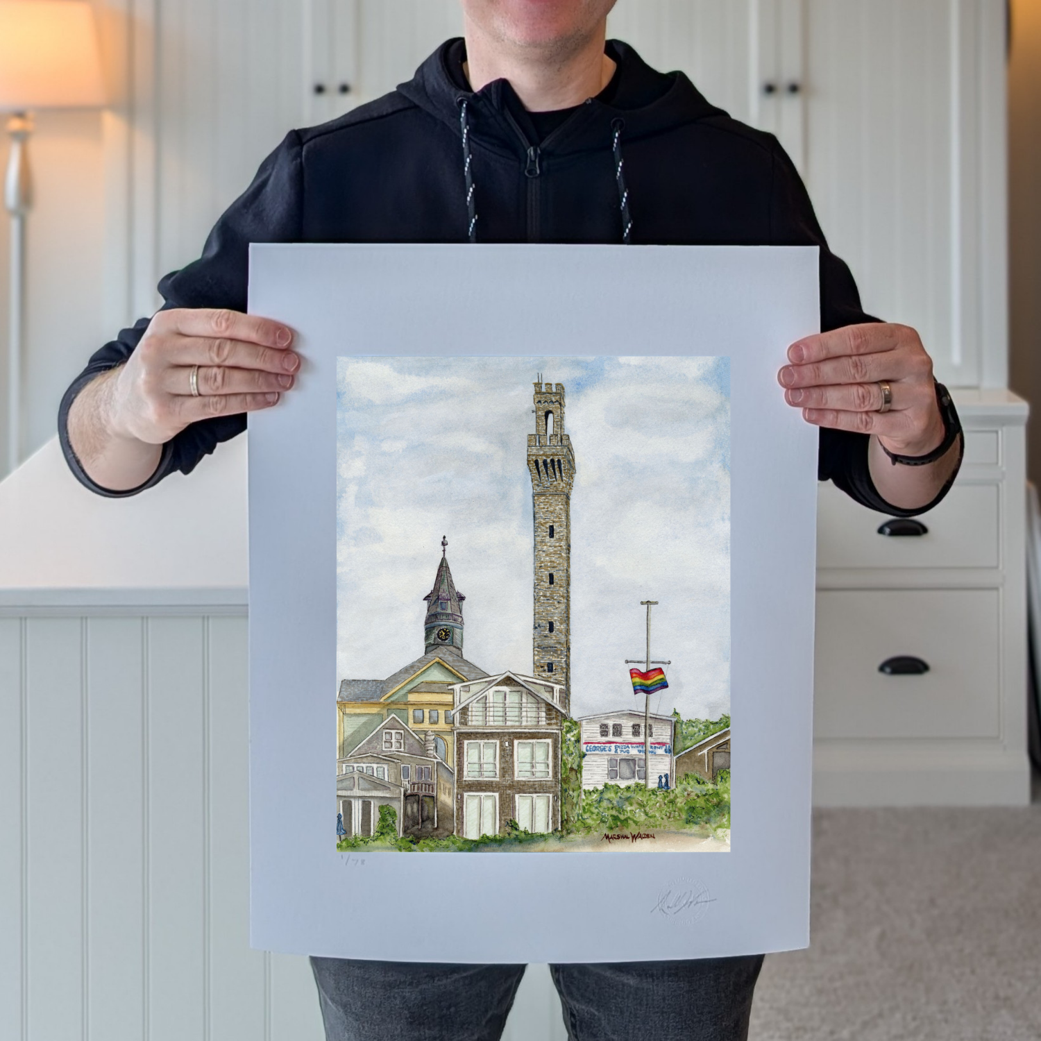 A photograph of artist Marshal Walden holding the large, 16x20 inch limited edition fine art print "Monumental Pride." The image demonstrates the scale of the artwork relative to the artist and highlights the detailed watercolor texture of the Provincetown skyline and the extra-wide crisp white borders. These borders allow for versatile framing, either presented without a mat for a modern look or matted to a standard 11x14 opening for a traditional presentation.
