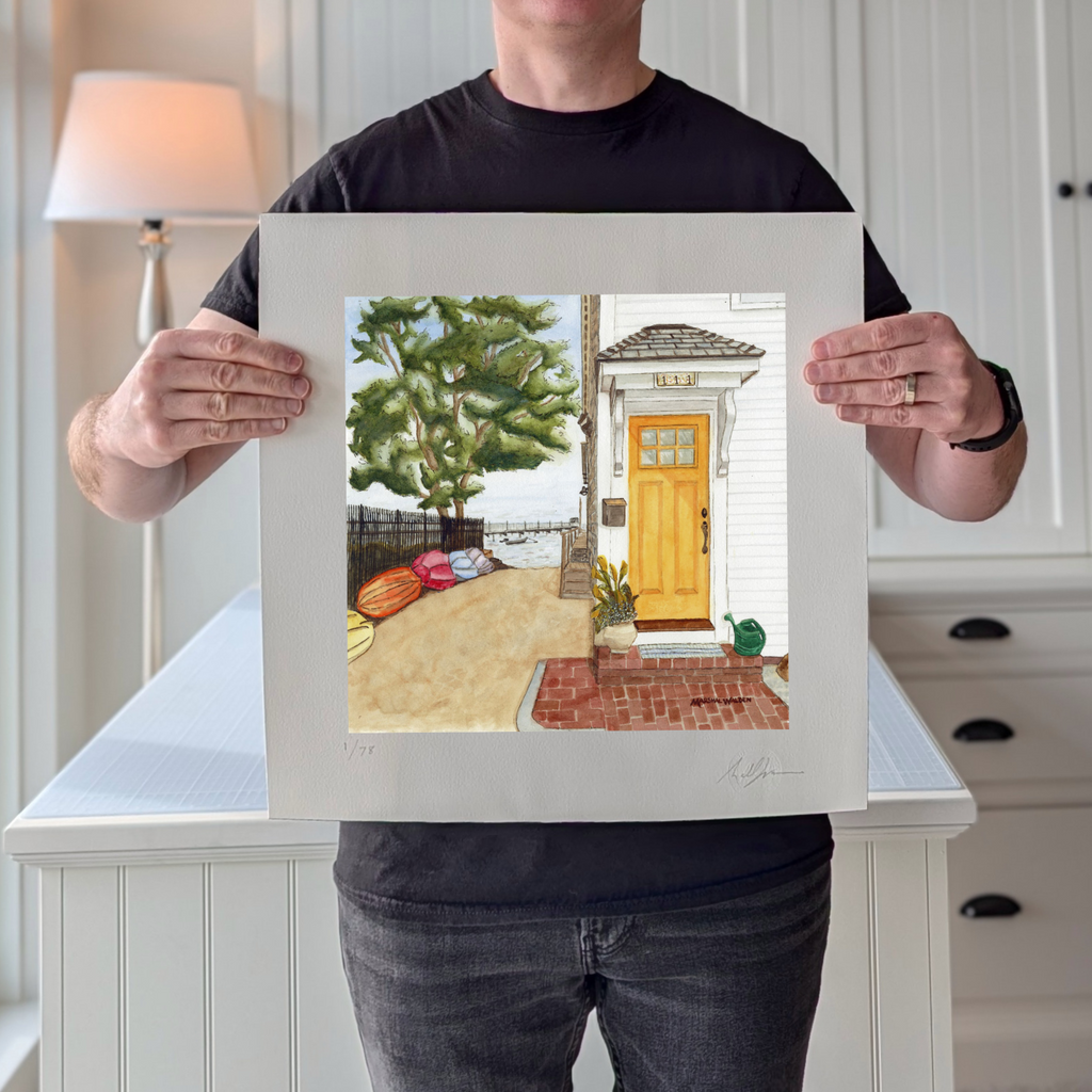 A photograph of PAAM member artist Marshal Walden holding the limited edition fine art print "The Turn - Yellow Door." The image demonstrates the scale of the artwork relative to the artist and highlights the detailed watercolor texture of the inviting bright yellow door, colorful kayaks, and sandy beach path leading to the harbor. The extra-wide crisp white borders are visible, allowing for versatile framing, either presented without a mat for a modern look or matted to a standard 10x10 opening for a tradi