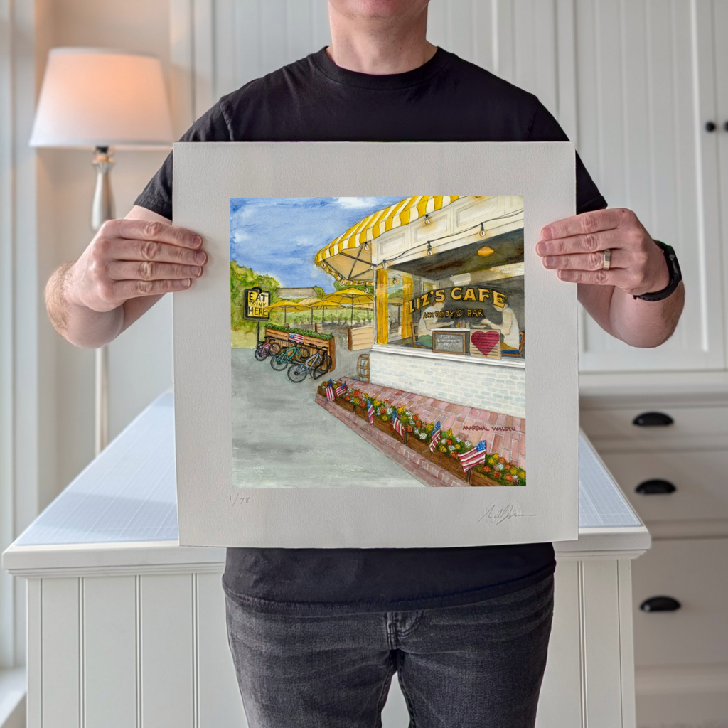 A photograph of artist Marshal Walden holding the limited edition fine art print "Window With A View." The image demonstrates the scale of the artwork relative to the artist and highlights the detailed watercolor texture of Liz's Café in Provincetown, featuring its iconic yellow umbrellas. The extra-wide crisp white borders are visible, allowing for versatile framing, either presented without a mat for a modern look or matted to a standard 10x10 opening for a traditional presentation.
