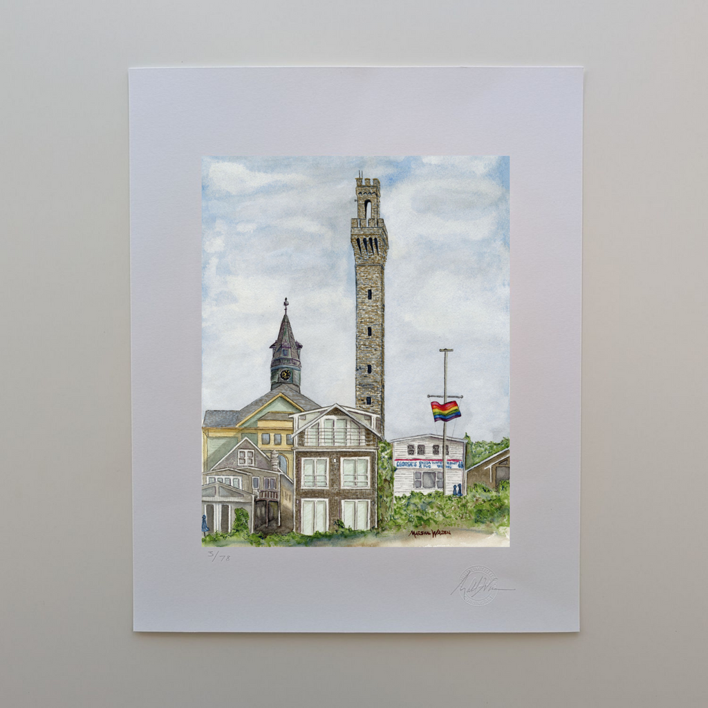 A photograph of a 16x20 limited edition fine art print titled "Monumental Pride" by artist Marshal Walden, resting on an off-white tabletop. The watercolor artwork depicts the historic Provincetown skyline with the tall granite Pilgrim Monument and the town hall clock tower rising above local homes and businesses. A rainbow Pride flag waves prominently on a flagpole to the right. The print's wide white border features the artist's hand-penciled signature, the edition numbering out of 78, and the official "C