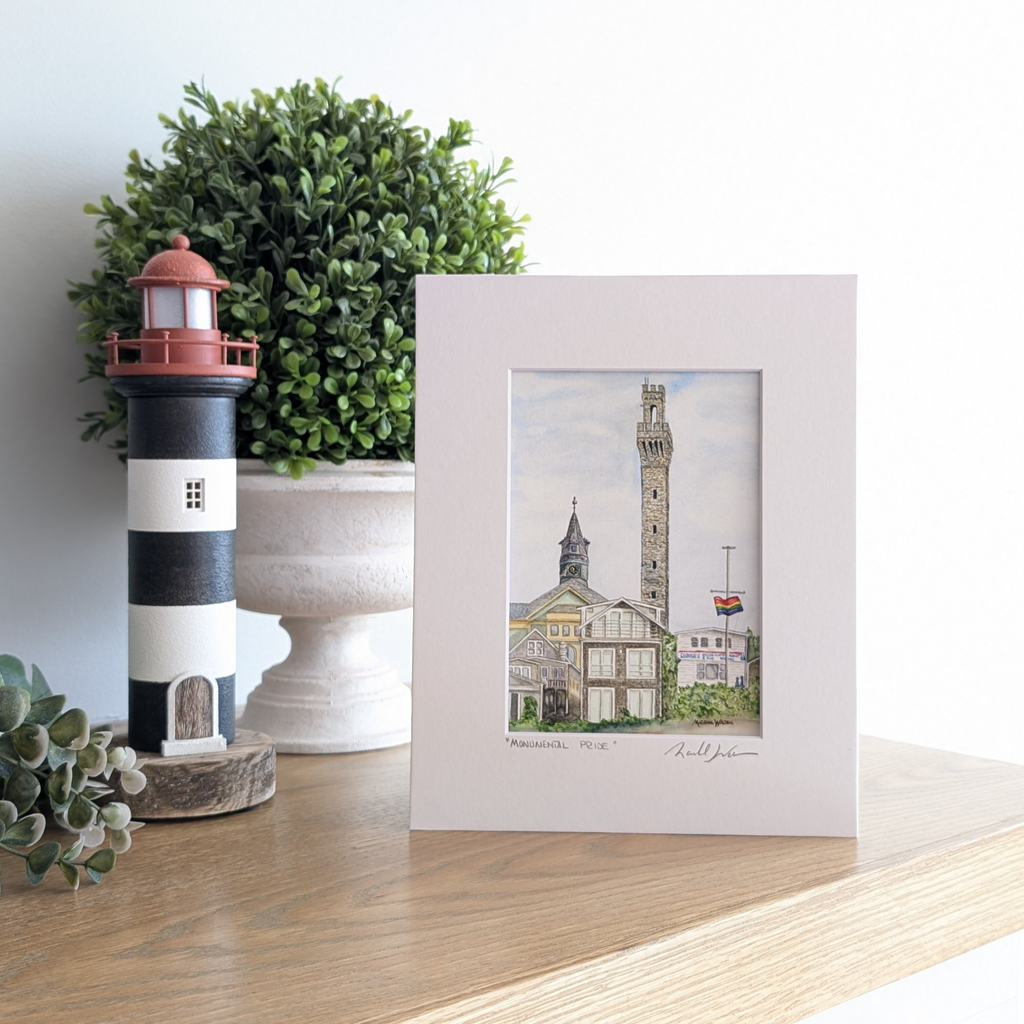 A lifestyle photograph of a professionally printed watercolor print titled "Monumental Pride" by PAAM artist Marshal Walden, propped up on a wooden mantel. The artwork depicts the Provincetown skyline with the tall granite Pilgrim Monument soaring above the town hall clock tower and historic coastal buildings. The print sits on a mantel next to a black and white striped lighthouse figurine and a lush green plant.
