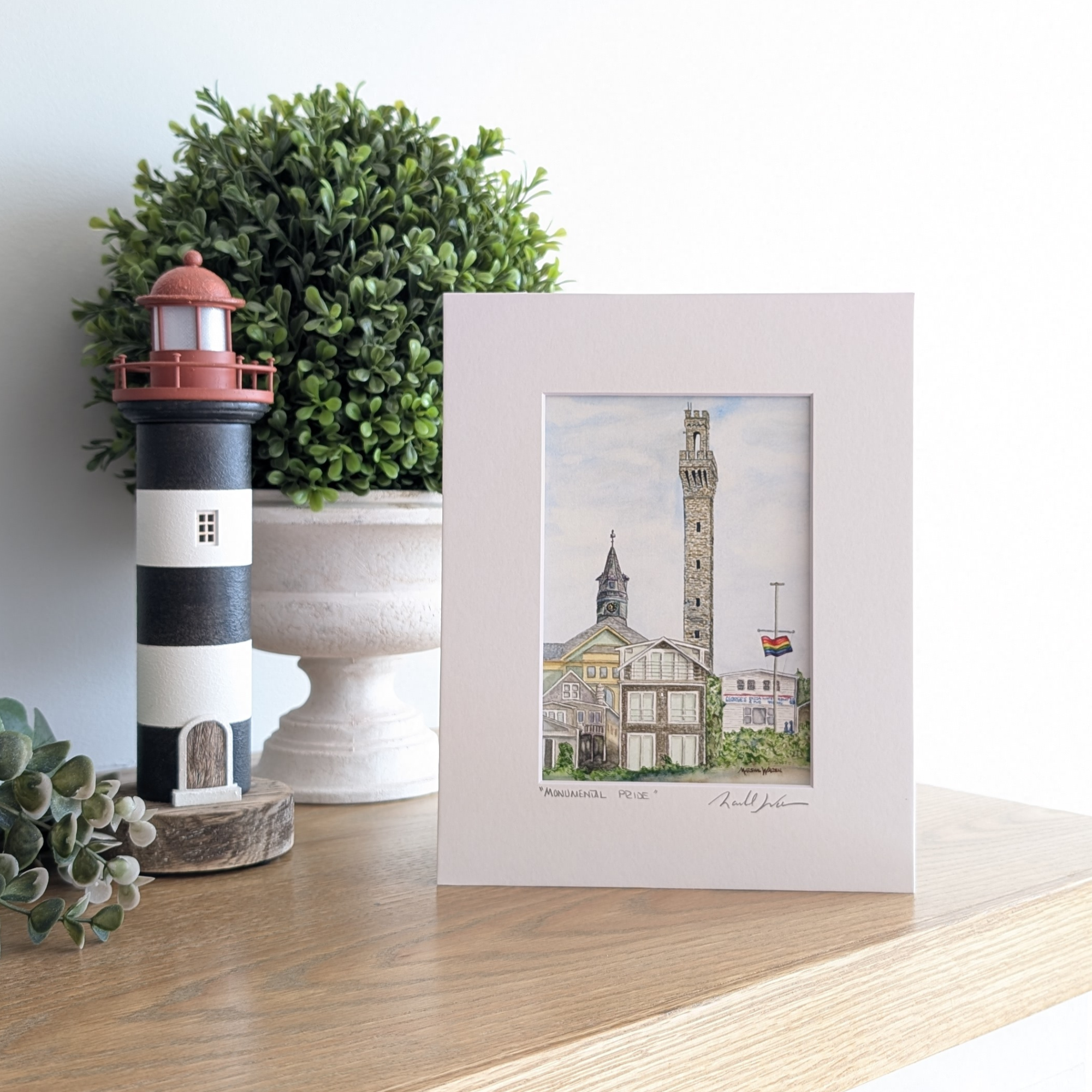 A lifestyle photograph of a professionally printed watercolor print titled "Monumental Pride" by PAAM artist Marshal Walden, propped up on a wooden mantel. The artwork depicts the Provincetown skyline with the tall granite Pilgrim Monument soaring above the town hall clock tower and historic coastal buildings. The print sits on a mantel next to a black and white striped lighthouse figurine and a lush green plant.
