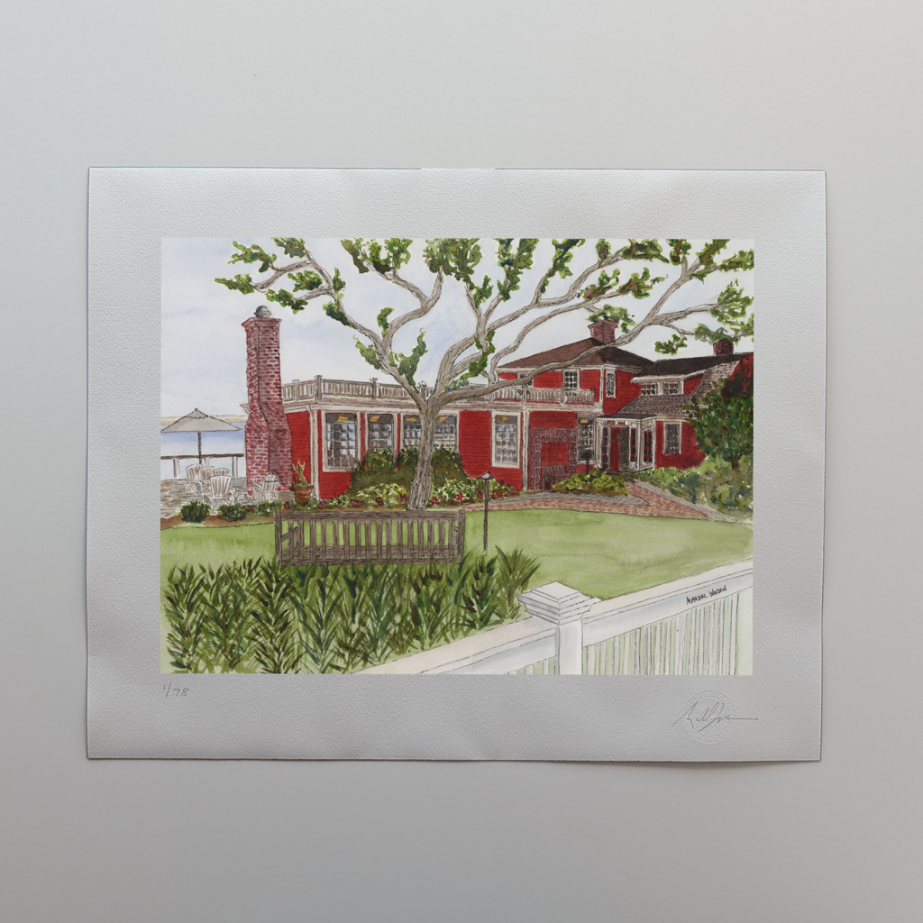 A photograph of a 16x20 limited edition fine art print titled "Red Inn" by PAAM member artist Marshal Walden, resting on an off-white tabletop. The watercolor artwork depicts the historic Red Inn in Provincetown, featuring its red architecture, lush gardens, and a welcoming deck with Adirondack chairs overlooking the harbor. The white border displays the artist's hand-penciled signature, the edition number out of 78, and the raised "Curbing Art" embossed seal.
