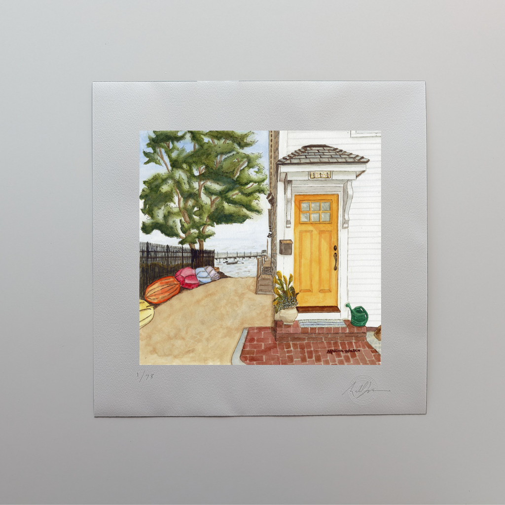 A photograph of a 16x16 limited edition fine art print titled "The Turn - Yellow Door" by PAAM member artist Marshal Walden, resting on an off-white tabletop. The watercolor artwork depicts a bright yellow door on a two-story cottage along Commercial Street in Provincetown, leading the eye past colorful kayaks and down a sandy beach path toward the harbor. The print’s wide white border displays the artist's hand-penciled signature, the edition numbering out of 78, and the official "Curbing Art" embossed sea
