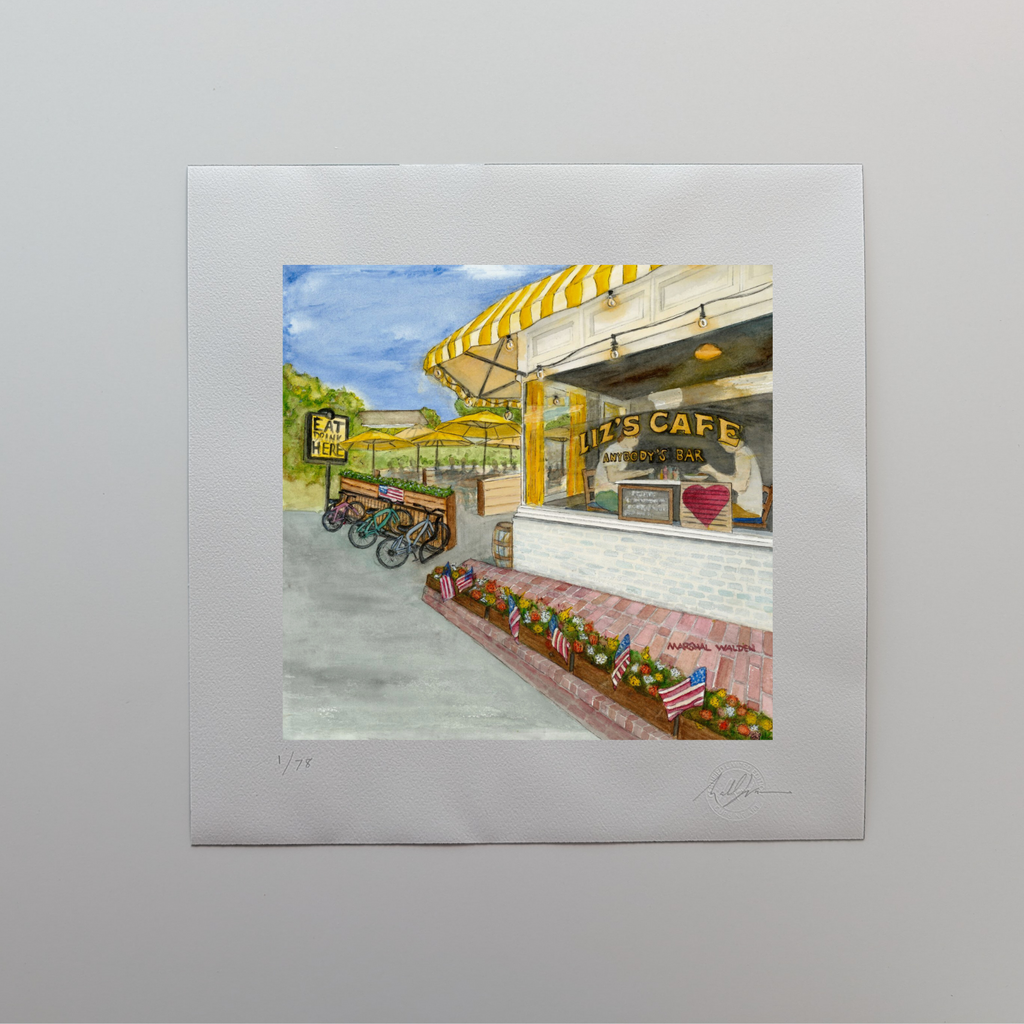 A photograph of a 16x16 limited edition fine art print titled "Window With A View" by PAAM member artist Marshal Walden, resting on an off-white tabletop. The watercolor artwork depicts Liz's Café (Anybody's Bar) on Bradford Street in Provincetown, featuring iconic bright yellow umbrellas, flower boxes, and large picture windows. The print’s wide white border displays the artist's hand-penciled signature, the edition numbering out of 78, and the official "Curbing Art" embossed seal in the bottom right corne