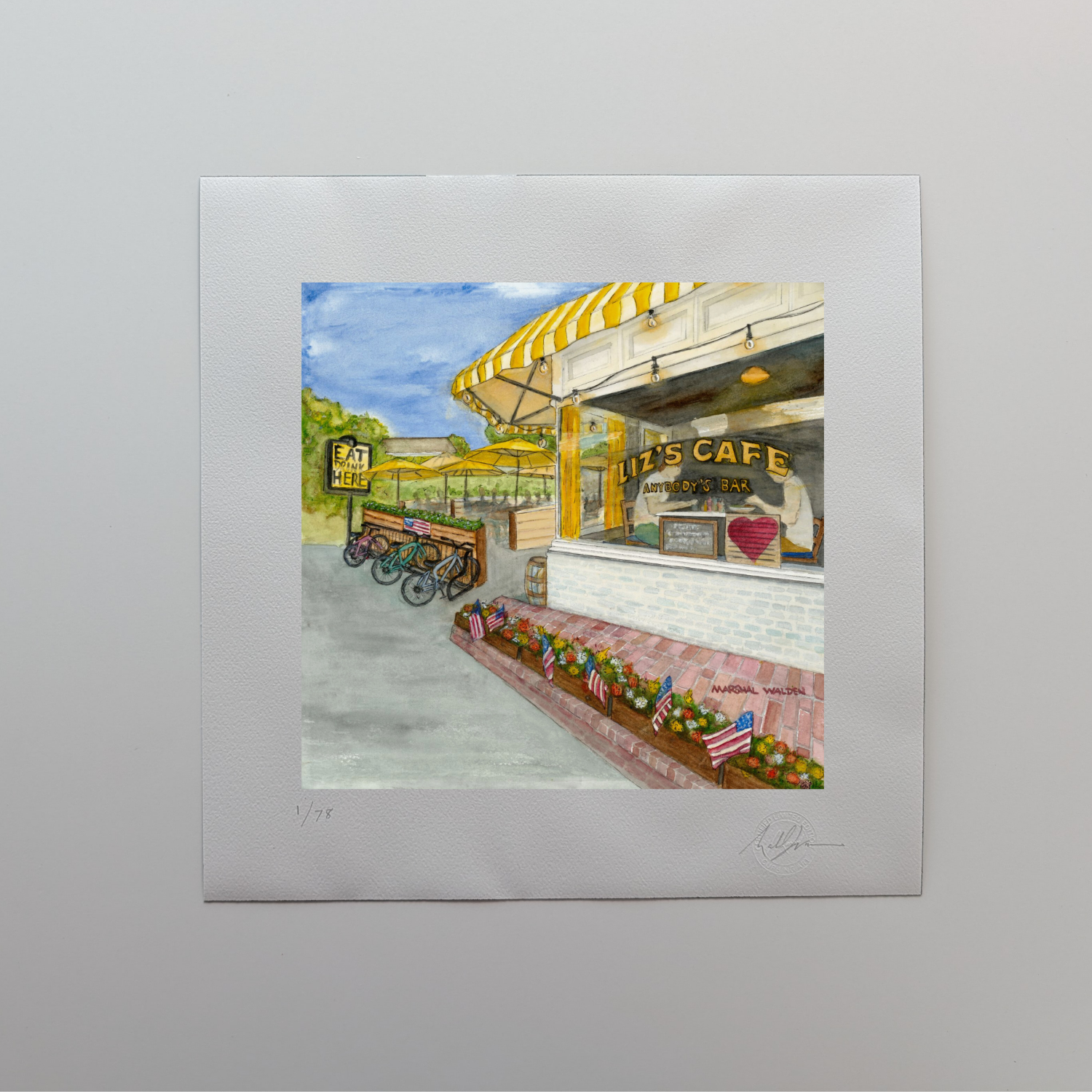 A photograph of a 16x16 limited edition fine art print titled "Window With A View" by PAAM member artist Marshal Walden, resting on an off-white tabletop. The watercolor artwork depicts Liz's Café (Anybody's Bar) on Bradford Street in Provincetown, featuring iconic bright yellow umbrellas, flower boxes, and large picture windows. The print’s wide white border displays the artist's hand-penciled signature, the edition numbering out of 78, and the official "Curbing Art" embossed seal in the bottom right corne